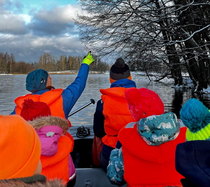 Viis noort ja kaks täiskasvanut on kummipaadis järvel. puud ja maa on lumine. Järvel vesi lainetab. Inimestel on seljas oranžid päästevestid.